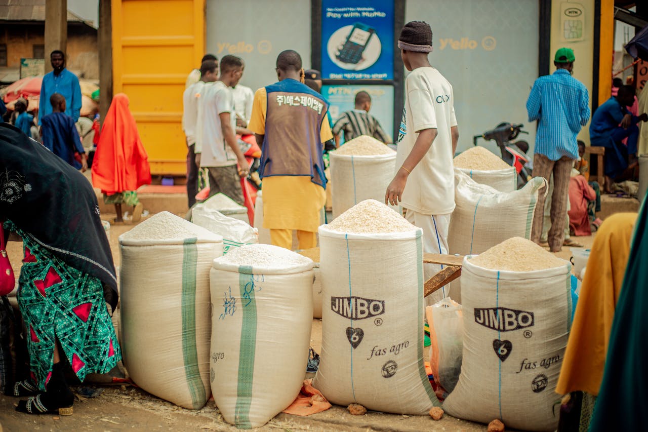 Bustling outdoor market with people and large rice sacks, vibrant community setting.